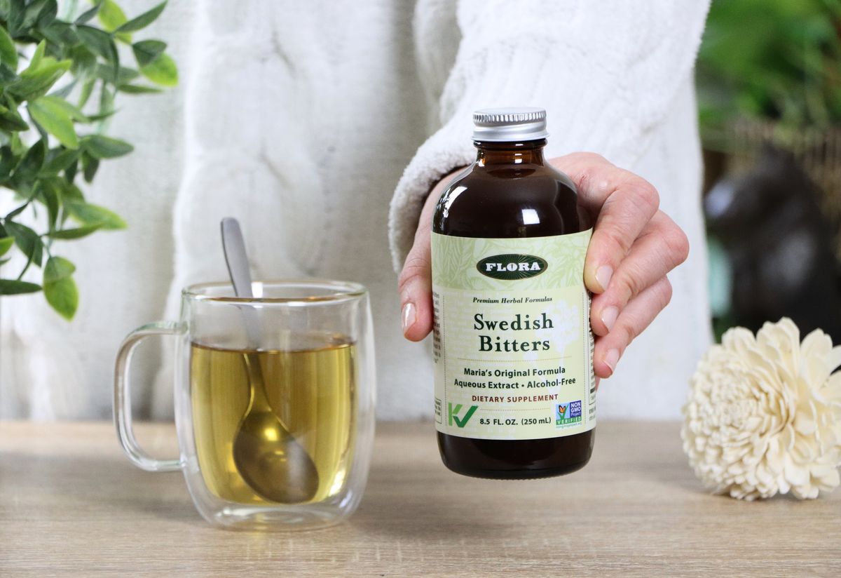 A woman holding a bottle of Swedish Bitters from Flora next to a cup of tea. 