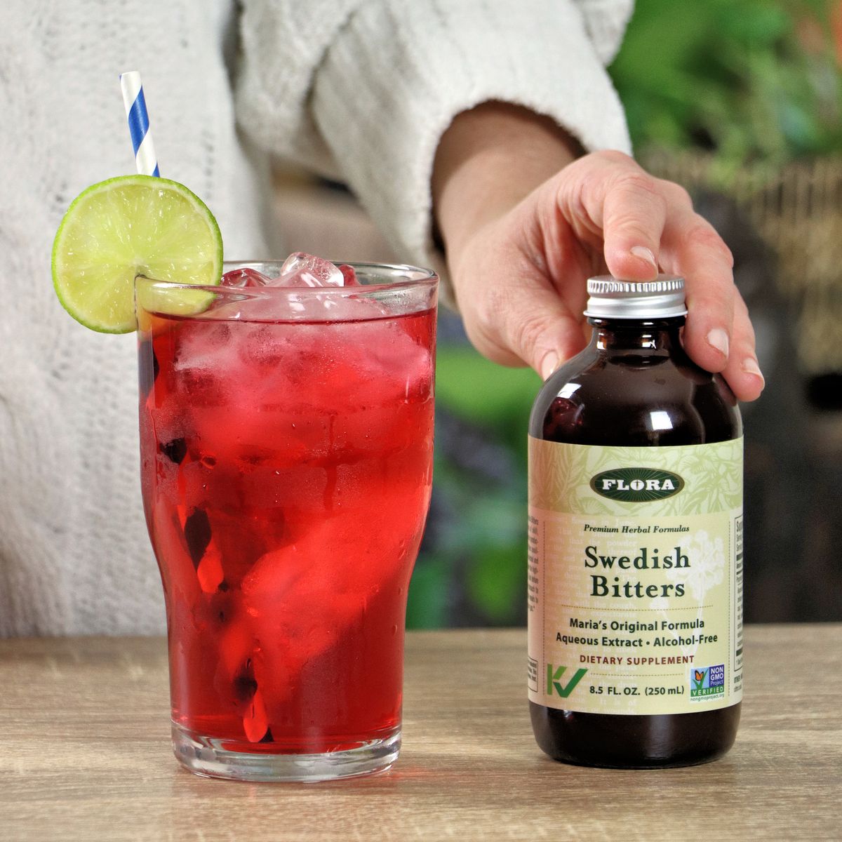 A woman holding a bottle of Swedish Bitters next to a chilled glass of hibiscus tea. 