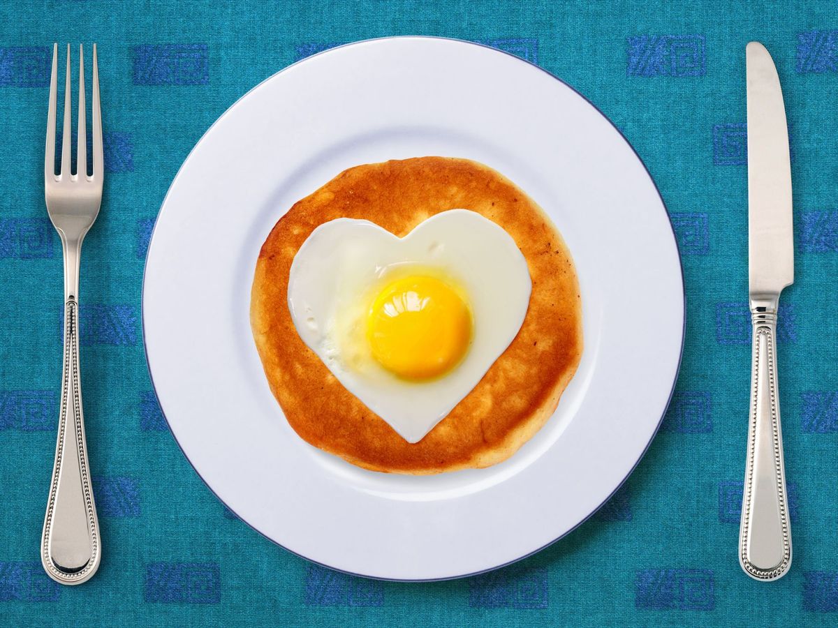 Ein herzförmiges Ei auf einem Pfannkuchen auf einem Teller neben Messer und Gabel.