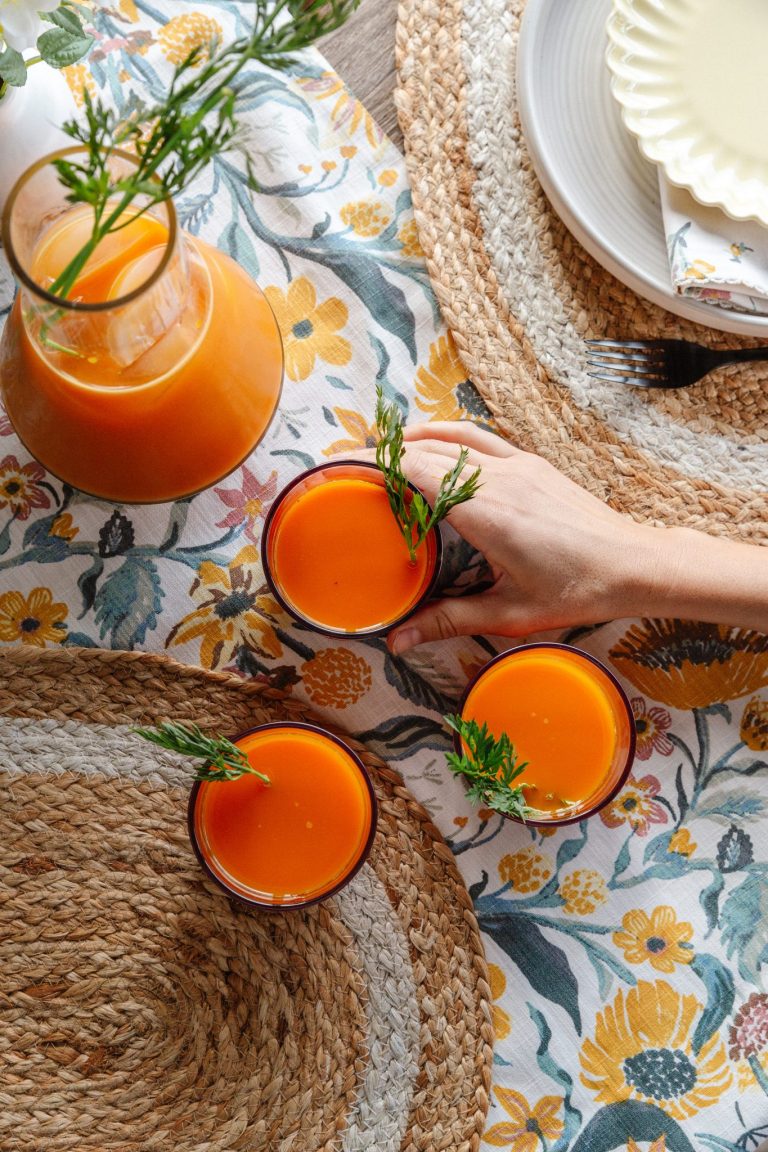 Drei Gläser Orangenkarottensaft auf dem Tisch mit gewebtem Tischset und geblümter Tischdecke.